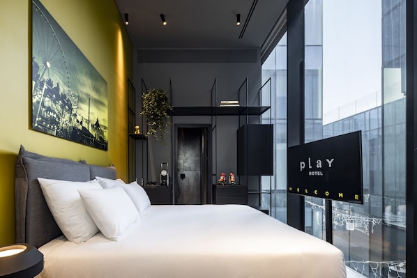 A modern hotel room with a large bed, a cityscape painting, a digital welcome sign, and a view through large windows. - Play Midtown (Tel Aviv)