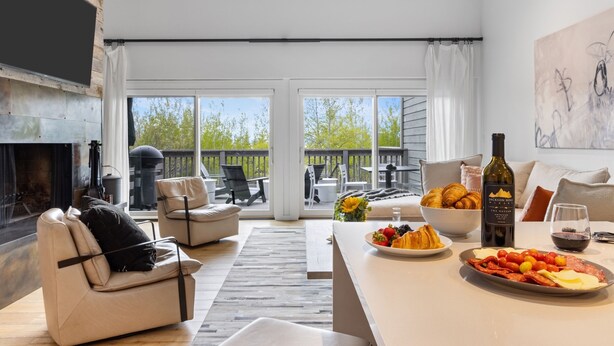 A modern living room with a fireplace, comfortable seating, a dining area with food and drinks, and a view of the outdoors. - Outpost: Timber Ridge 9 - Spacious Deck, Hot Tub & Pool Access! (Teton Village)