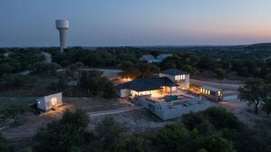 Een groot, modern huis met meerdere kamers en een zwembad, omgeven door bomen en een watertoren op de achtergrond.