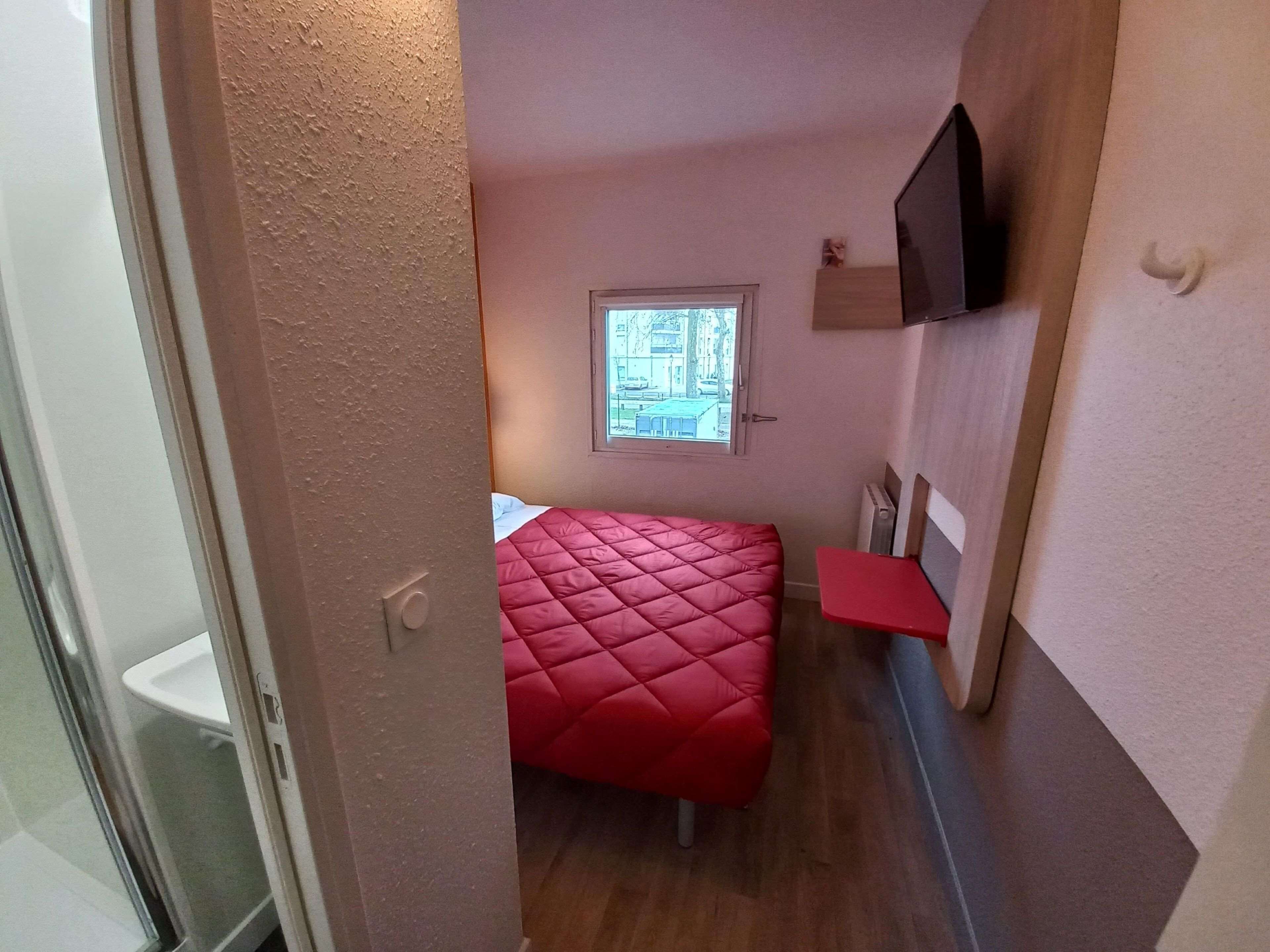 A small, compact room with a red bed, a window with a view of buildings, a mounted TV, and a small shelf.