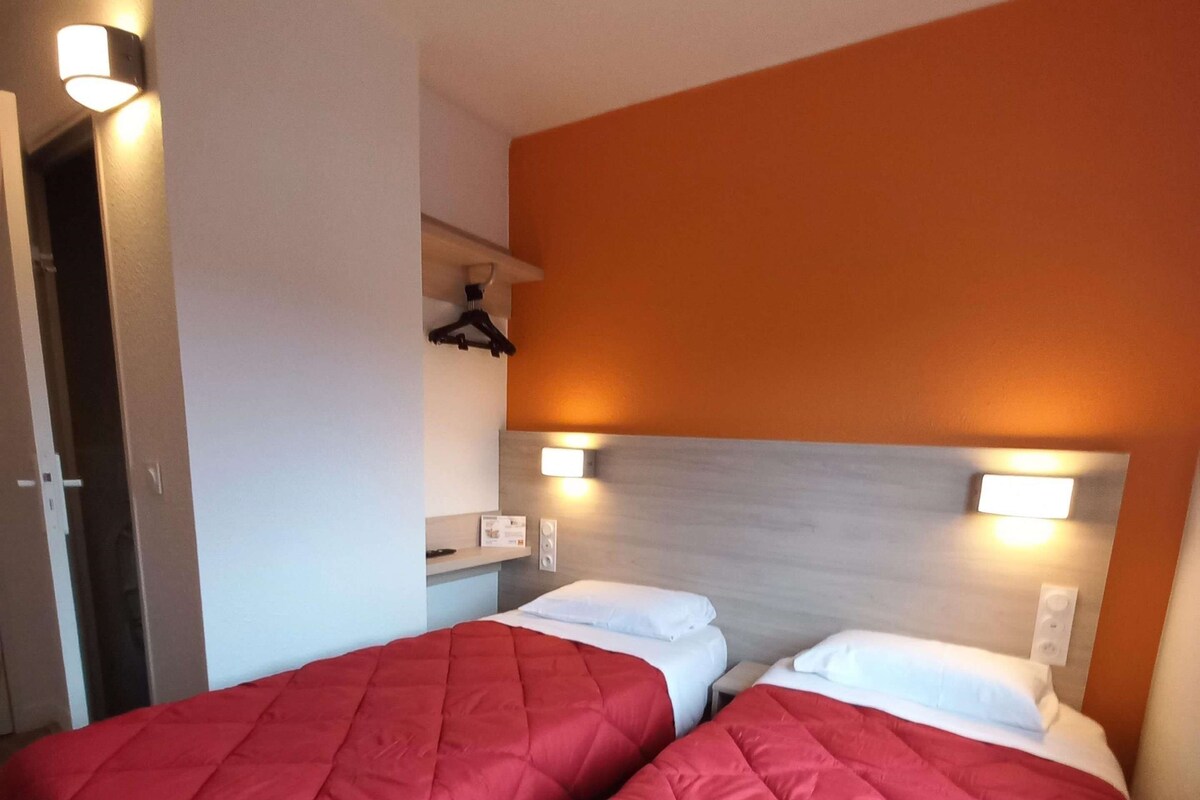 Une chambre d’hôtel avec deux lits, chacun recouvert d’une courtepointe rouge matelassée et agrémenté d’oreillers blancs. Des appliques murales sont fixées au-dessus de la tête de lit, et une petite étagère porte un miroir et un porte-manteau.