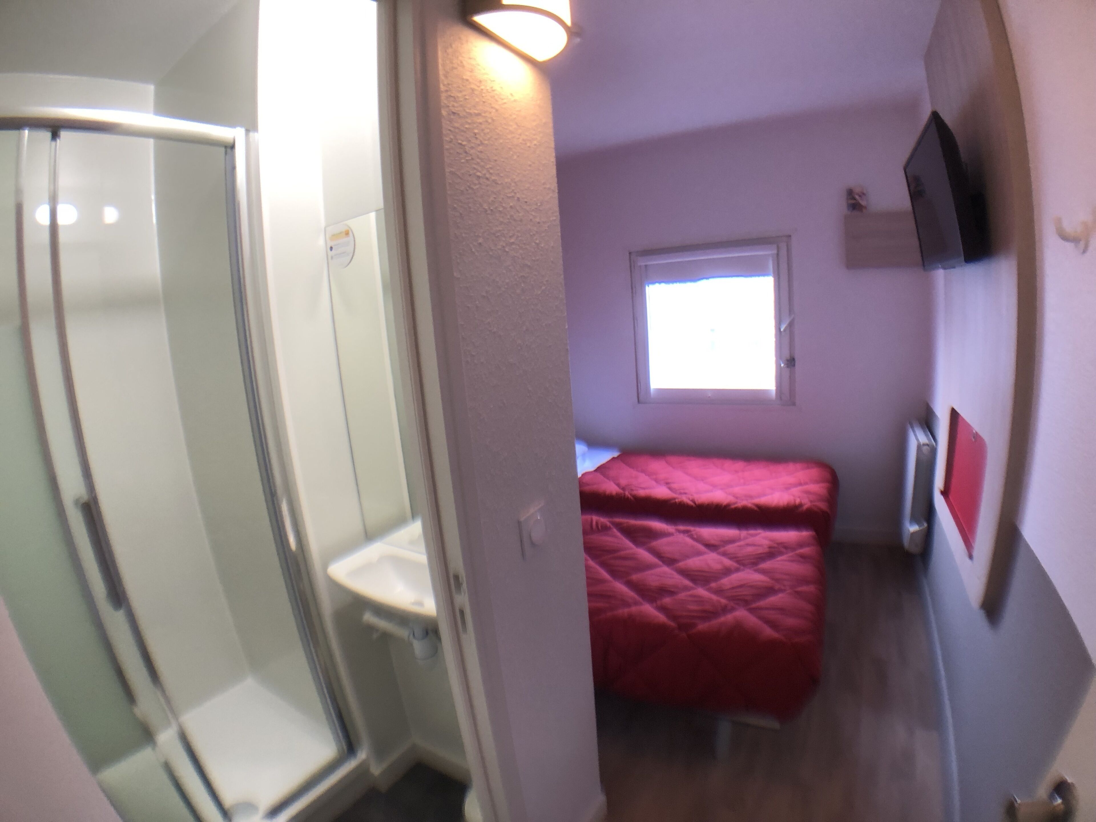 A compact room with a shower, a bed, a window, and a TV mounted on the wall.