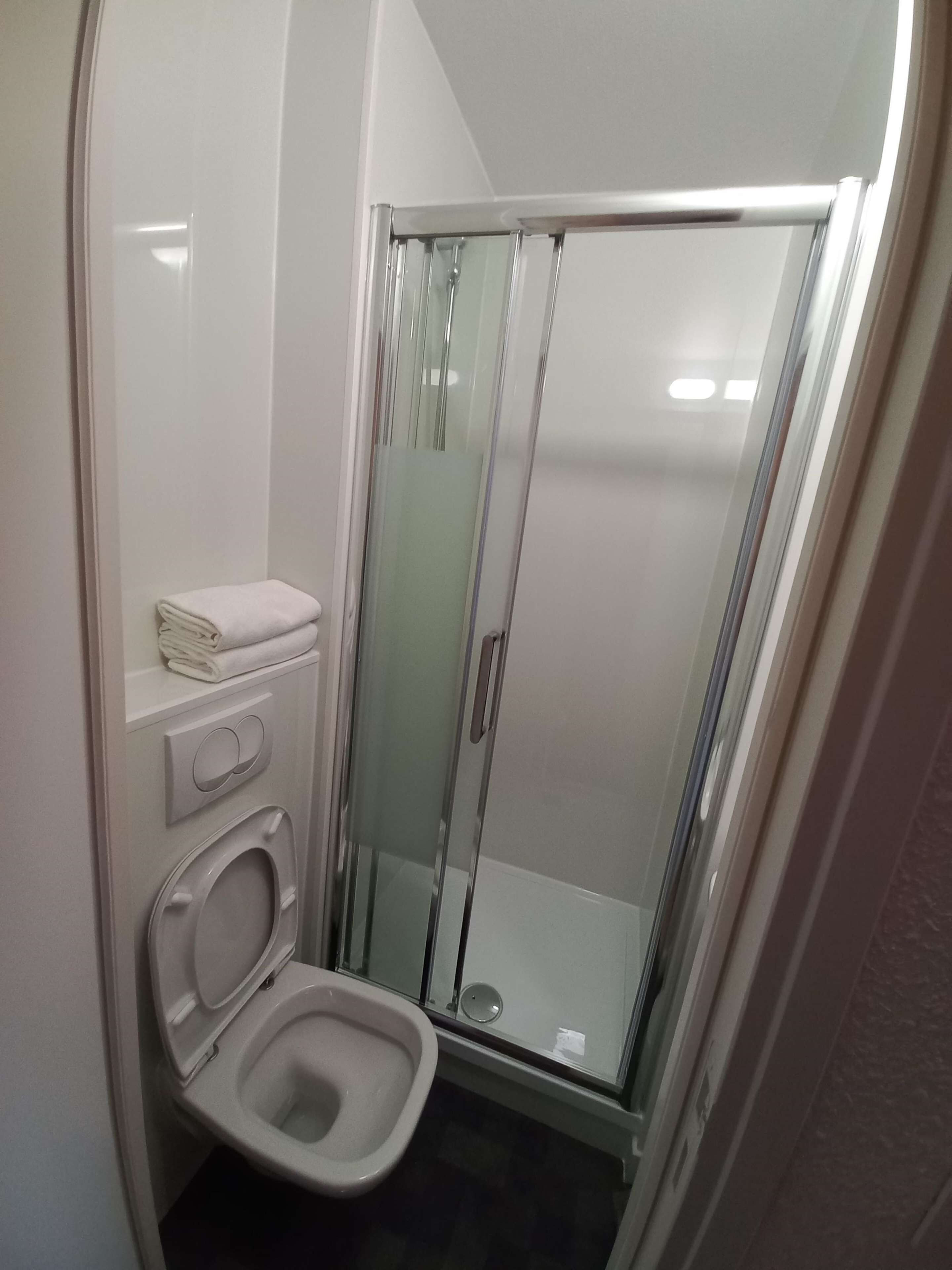 A small, compact bathroom with a glass-enclosed shower, a toilet, and a stack of folded towels on a shelf.