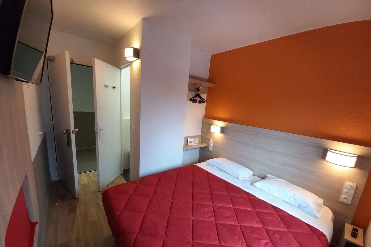 Une chambre d’hôtel comprenant un lit, une table de chevet, une lampe fixée au mur et une porte donnant sur une autre pièce.