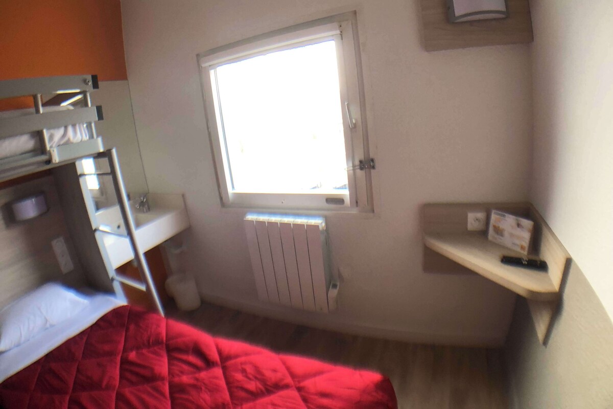 Une chambre de dortoir avec des lits superposés, une fenêtre, un radiateur et une petite étagère sur laquelle se trouve un livre.