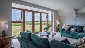 Ein geräumiges Wohnzimmer mit einem grünen Sofa, einem runden Couchtisch und Blick auf ein grünes Feld durch große Fenster.