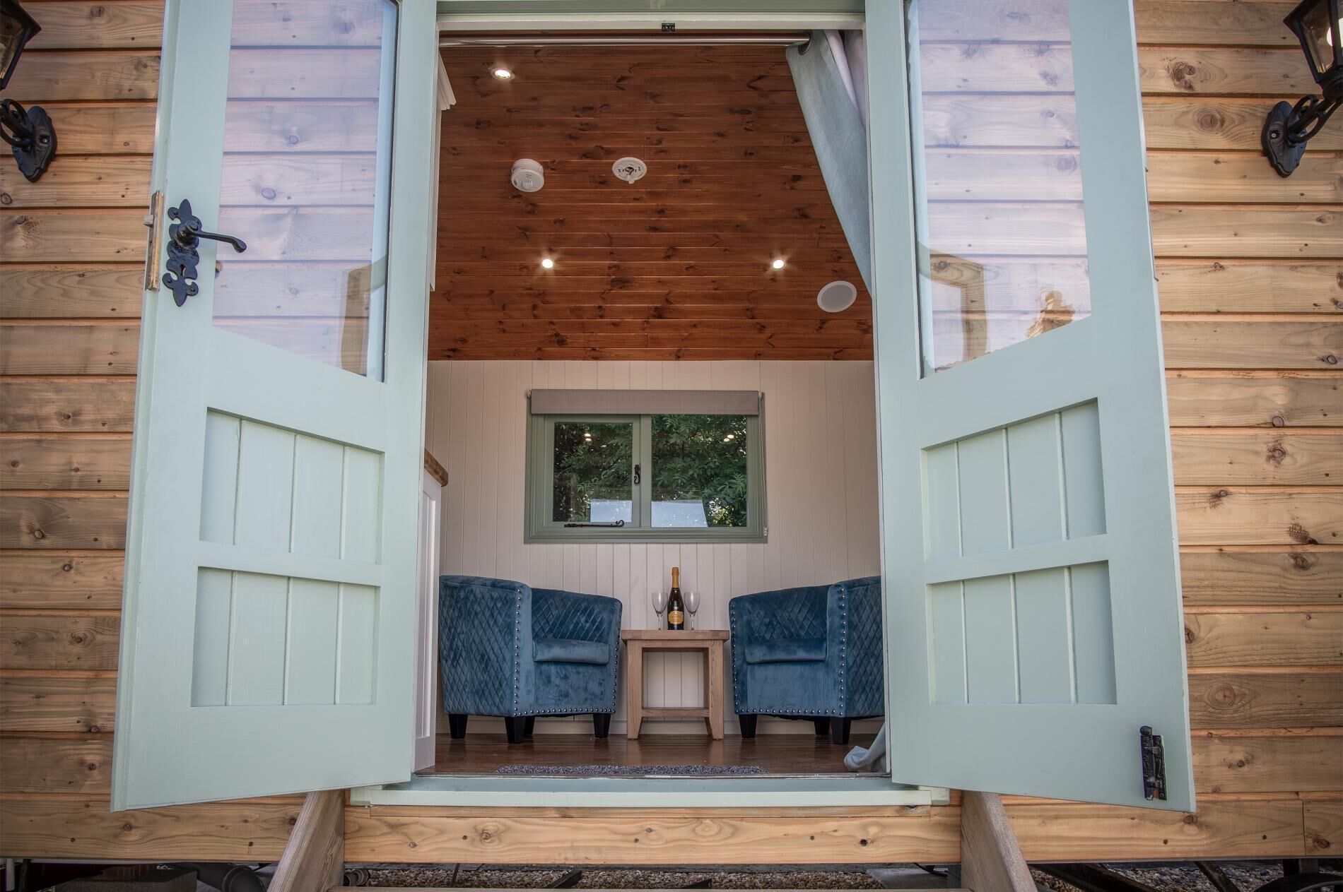 A wooden cabin with a large open door leading to a room with two blue upholstered chairs, a small table, and a window.