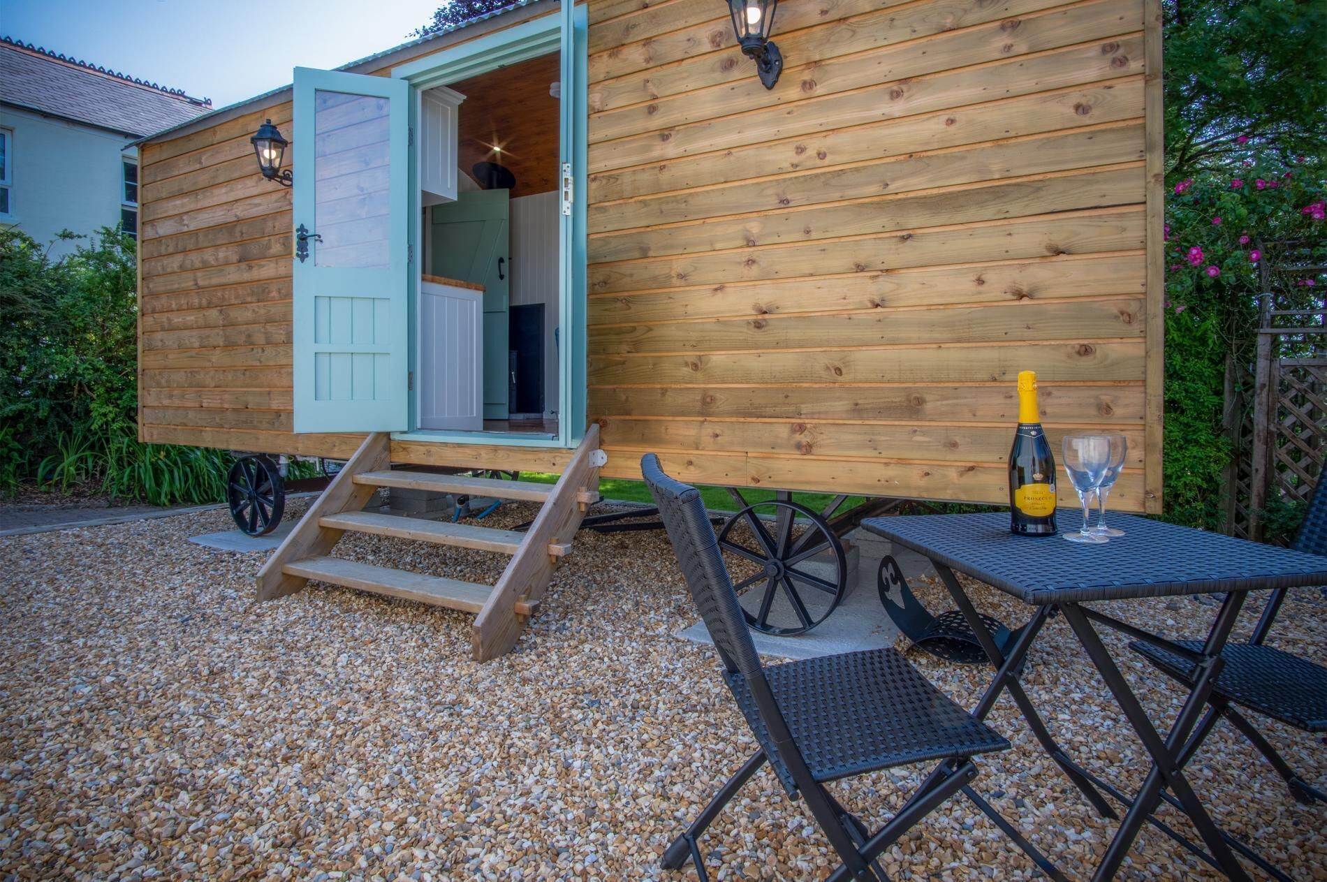 A wooden cabin with a door open, a table with a bottle and glass, and a chair.