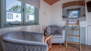 A cozy room with a large grey quilted chair, a small wooden table with a bottle and glasses, a grey armchair, and a wooden ladder. - Willow - 1 Bedroom Shepherd's Hut - Amroth (Narberth)
