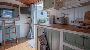 A cozy kitchen with wooden cabinets, a kettle, and a coffee maker. A ladder is leaning against the wall, and a window with curtains is visible. - Willow - 1 Bedroom Shepherd's Hut - Amroth (Narberth)