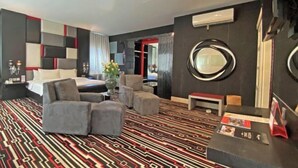 A modern hotel room with a patterned carpet, a bed with pillows, a seating area with chairs, a round mirror, and a small table with a magazine.