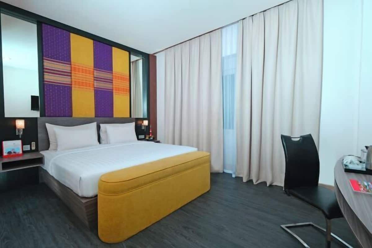 A hotel room with a large bed, a yellow ottoman, a desk with a chair, and a large window with curtains.