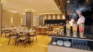 A restaurant with a chef preparing food, tables set with white tablecloths, and a buffet with various dishes and drinks.