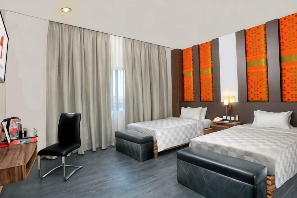 A hotel room with two beds, a desk with a chair, a small table with a lamp, and a large window with curtains.