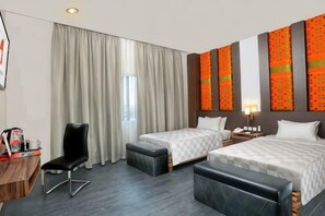 Ett hotellrum med två sängar, ett skrivbord med en stol, ett litet bord med en lampa och ett stort fönster med gardiner.