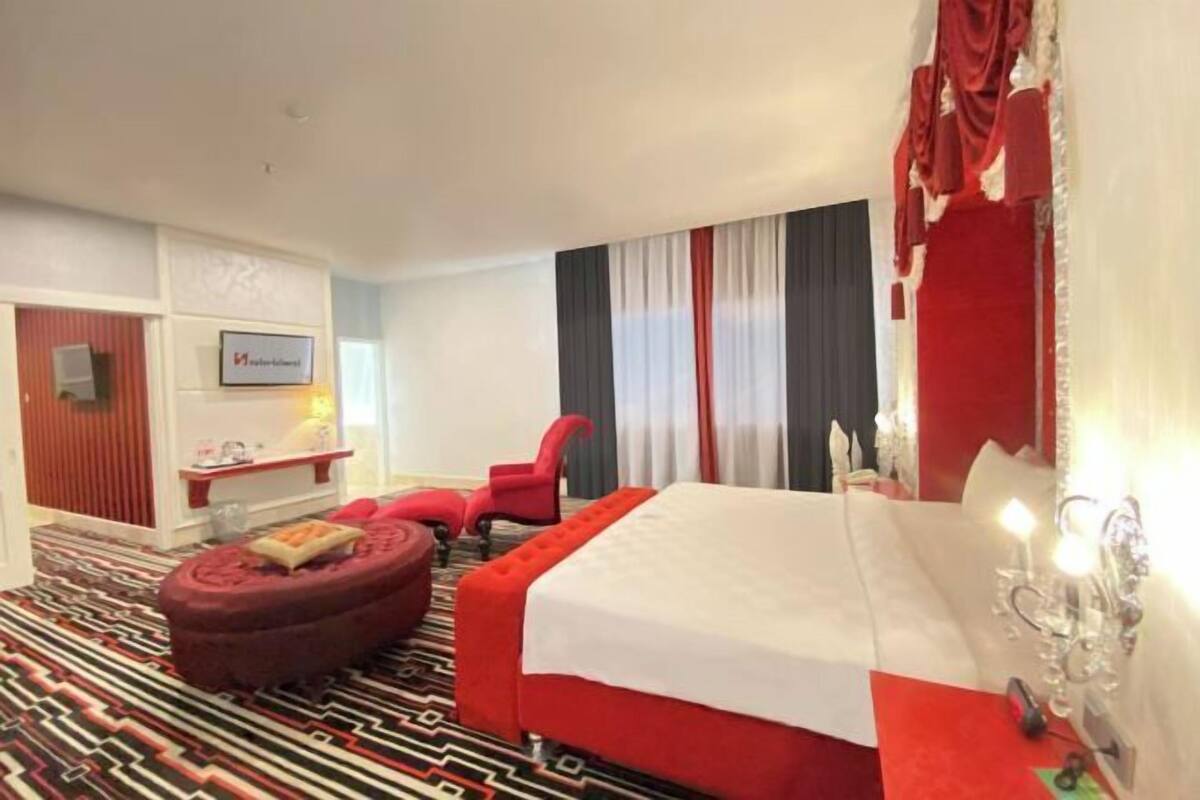 A hotel room with a bed, a red ottoman, a red chair, a desk with a lamp, and a wall-mounted TV.