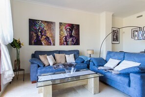 A living room with a blue sofa, a glass coffee table, and two paintings on the wall. - Charming apartment close to the beach. (Manilva)