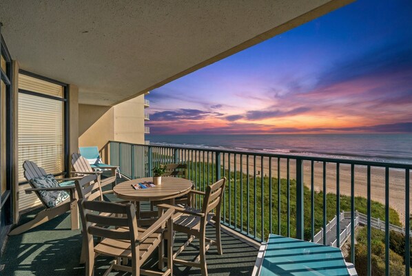 Un balcón con vista al océano al atardecer, muebles de madera y una pequeña mesa.