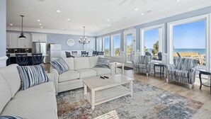 A spacious living room with a white sofa, a coffee table, and striped armchairs. There is a kitchen area with white cabinets and a refrigerator. Large windows offer a view of the ocean.
