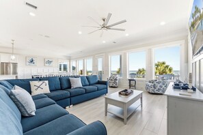 A spacious living room with a large blue sofa, a ceiling fan, and a view of the ocean through large windows.