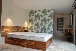 Une chambre moderne avec une tête de lit en bois, un grand lit, un mur recouvert d’un papier peint aux motifs tropicaux et un mur en pierre agrémenté d’une étagère intégrée.