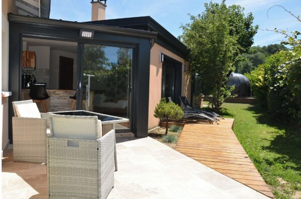 Une terrasse extérieure moderne, dotée de meubles en osier, d’un espace clos par des parois de verre et d’une plateforme en bois donnant sur un jardin avec une serre.