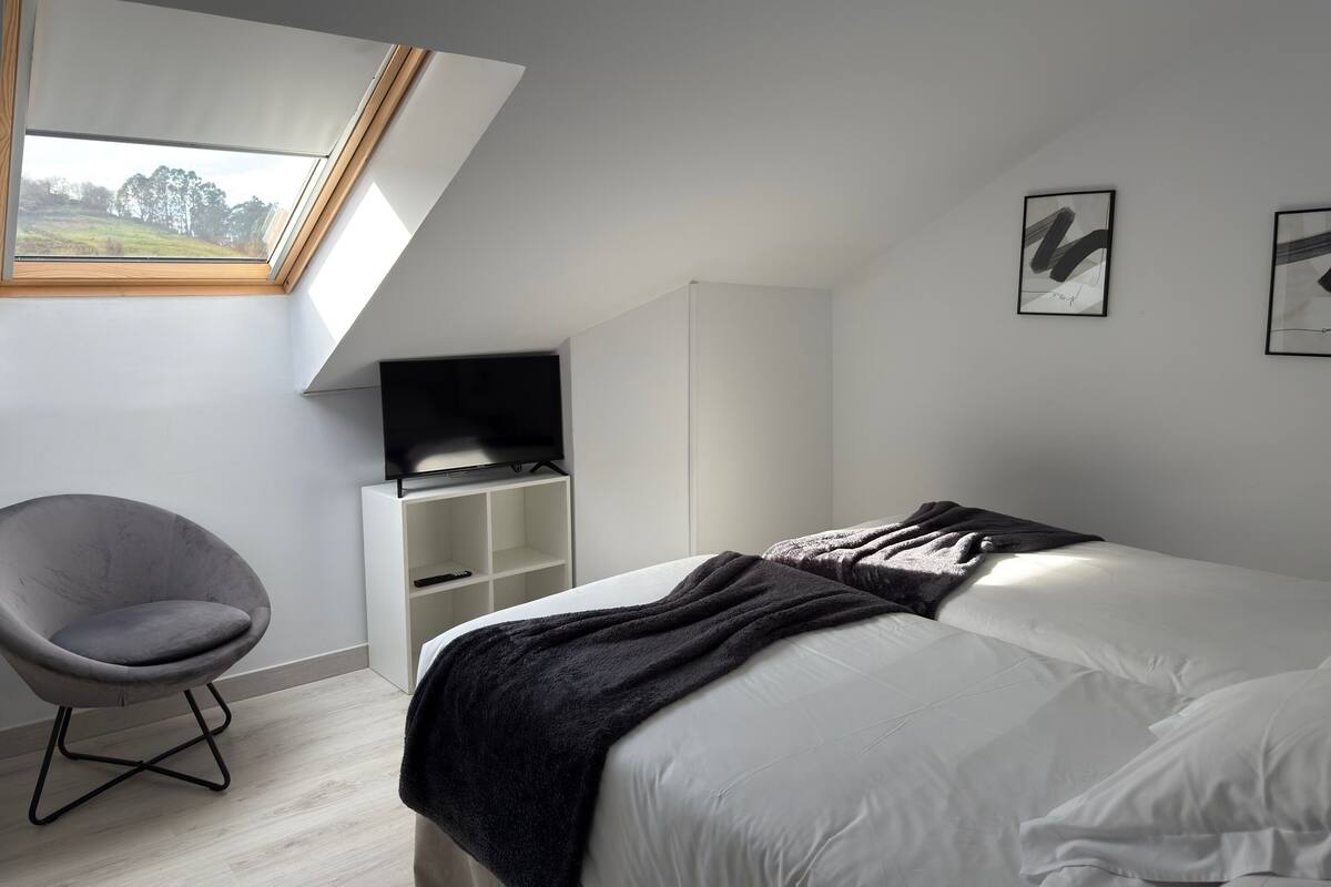 Een moderne slaapkamer met dakraam, een bed met donkere deken, een grijze stoel en een televisie op een witte dressoir.