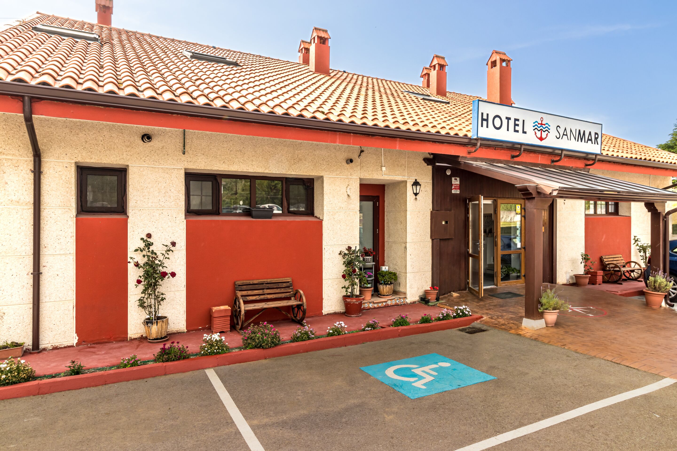 Un hotel con fachada roja y color crema, un letrero que dice "HOTEL SANMAR", y un espacio de estacionamiento accesible para sillas de ruedas.