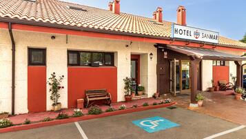 Un hotel con fachada roja y color crema, un letrero que dice "HOTEL SANMAR", y un espacio de estacionamiento accesible para sillas de ruedas.