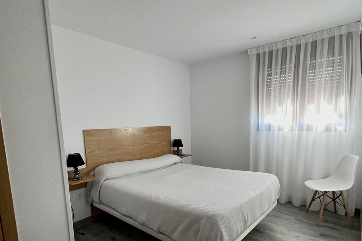Een slaapkamer met een bed met houten hoofdbord, een nachtkastje met lamp, een witte stoel en lichte gordijnen.