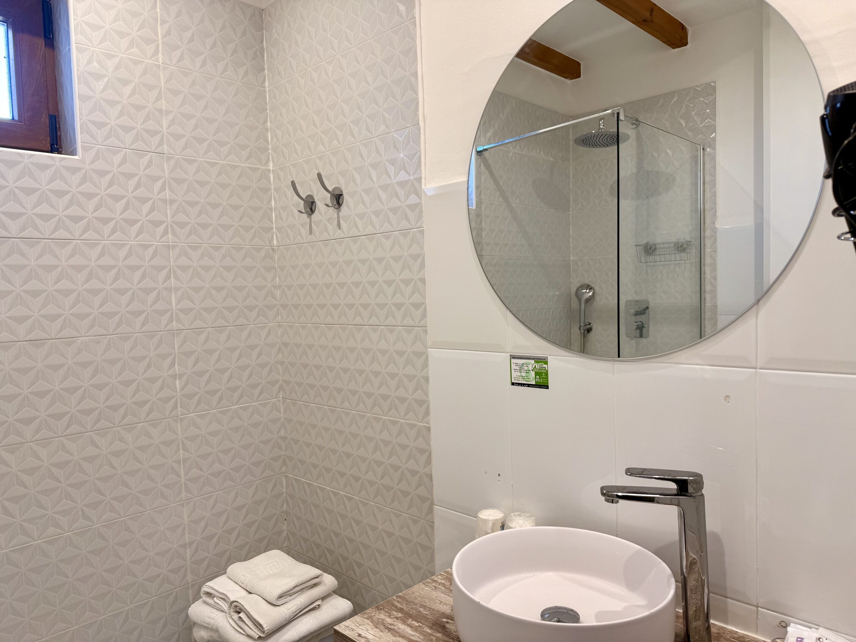 Un baño con pared de azulejos de diseño geométrico, ducha con cerramiento de vidrio, espejo redondo y lavabo con grifo.