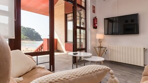 A room with a sofa, a TV, a small table, and a view of the beach. - Hotel Sanmar (Alfoz de Lloredo)