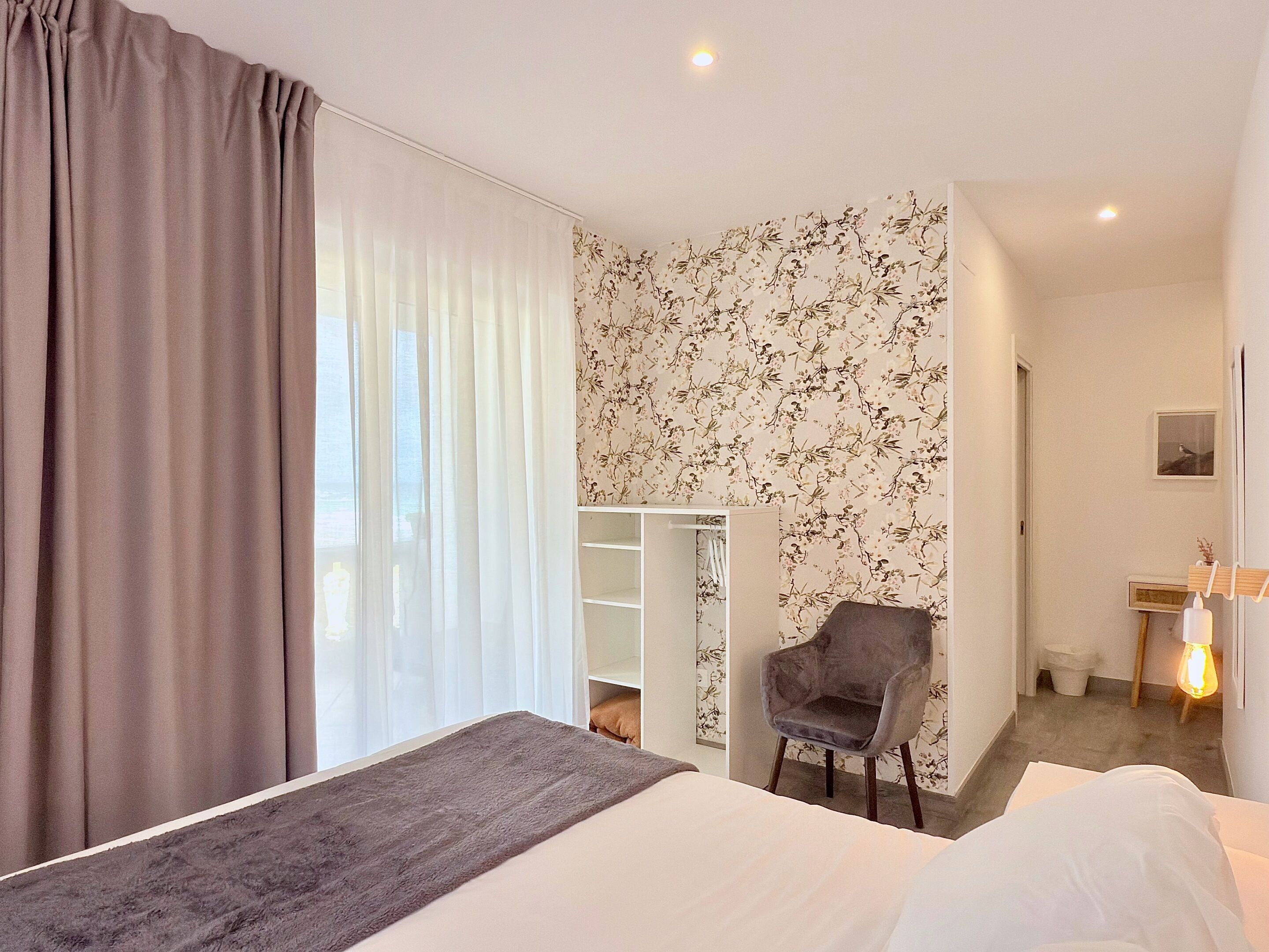 A modern hotel room with a large bed, a grey armchair, a wardrobe, and a floral wallpaper.