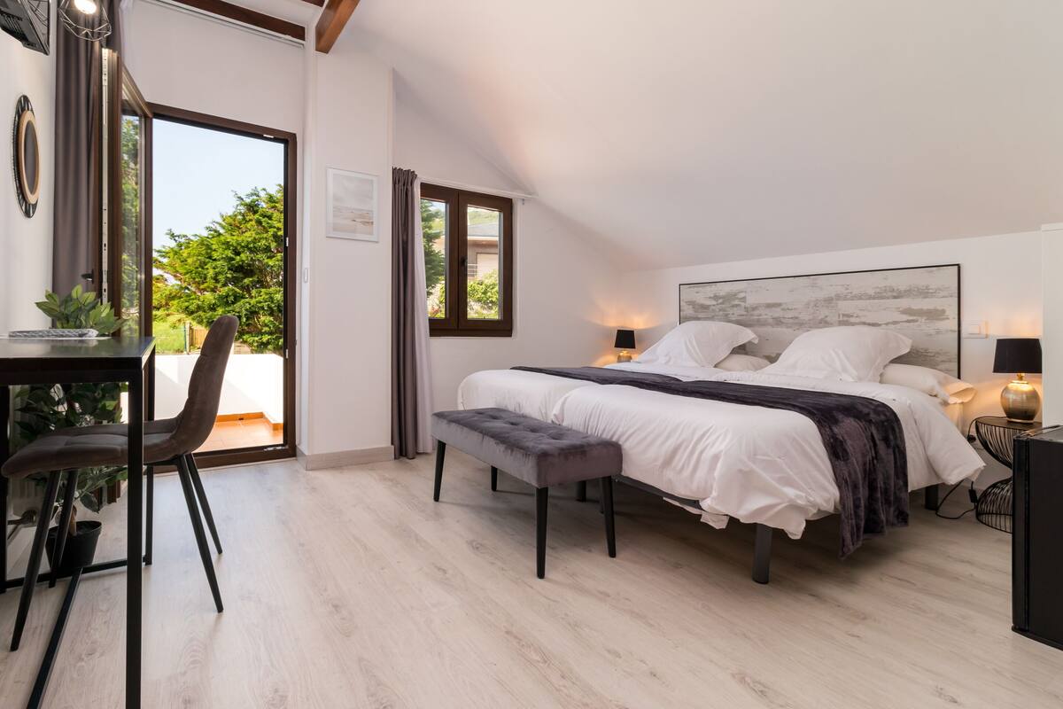 Een slaapkamer met een bed, een bankje, bureau, stoel en uitzicht op groen door een raam.
