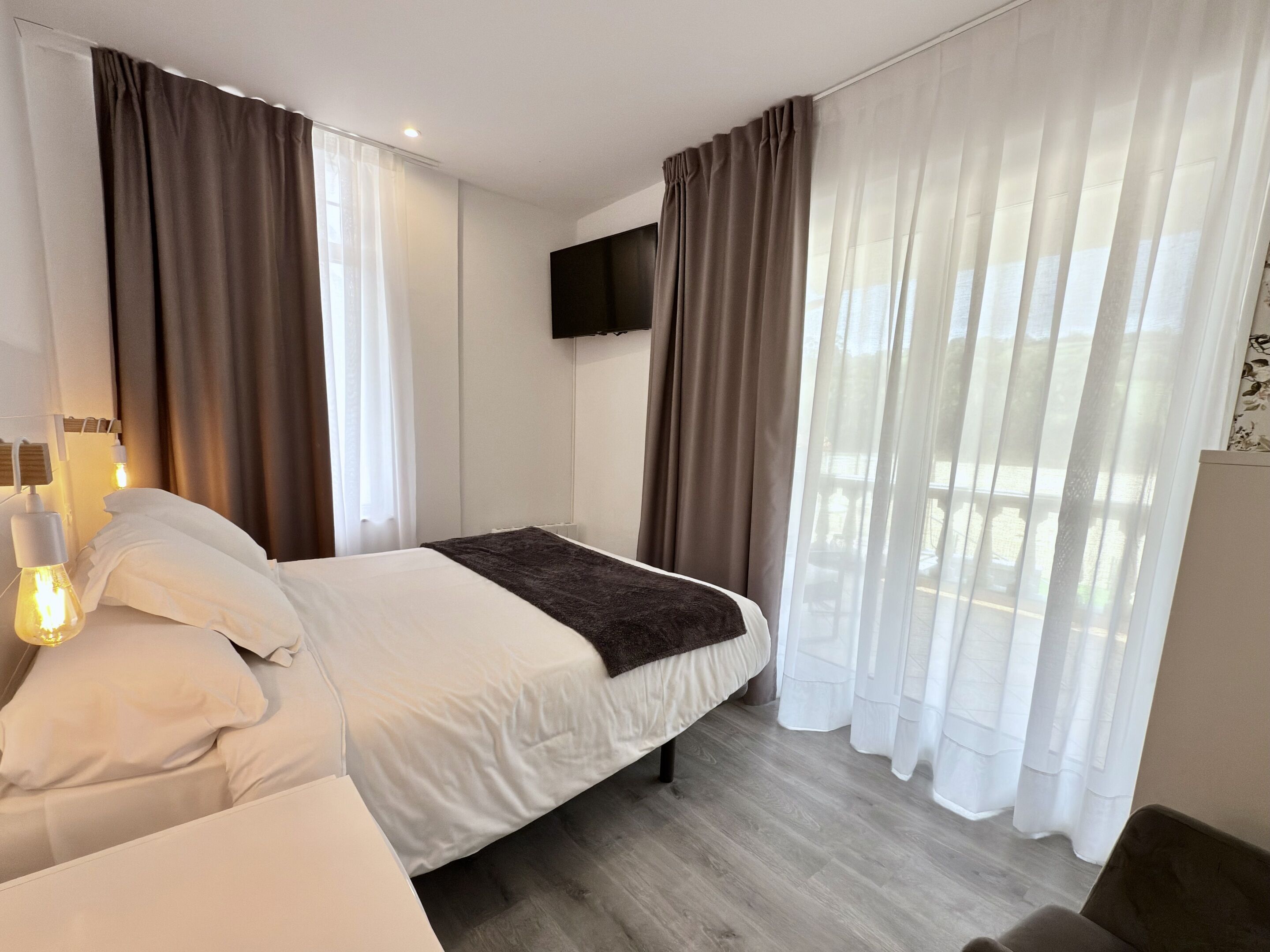 Una habitación de hotel moderna con cama, cortinas blancas, televisor montado y piso de madera.