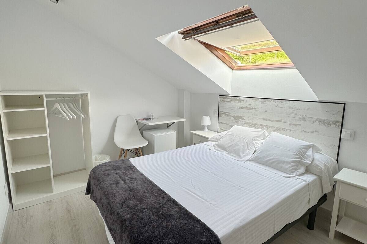 Een moderne slaapkamer met dakraam, een bed met witte lakens, een witte kledingkast en een bureau met stoel.