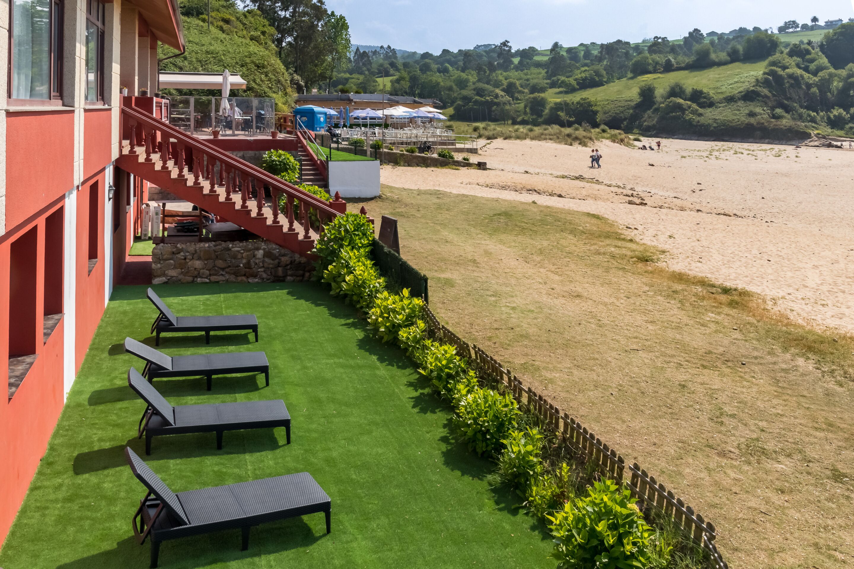Un hotel con fachada roja, una escalera que lleva a una terraza y vista al mar.