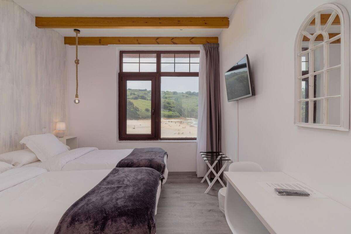 Een slaapkamer met twee bedden, een bureau, een televisie en een raam met uitzicht op het strand.