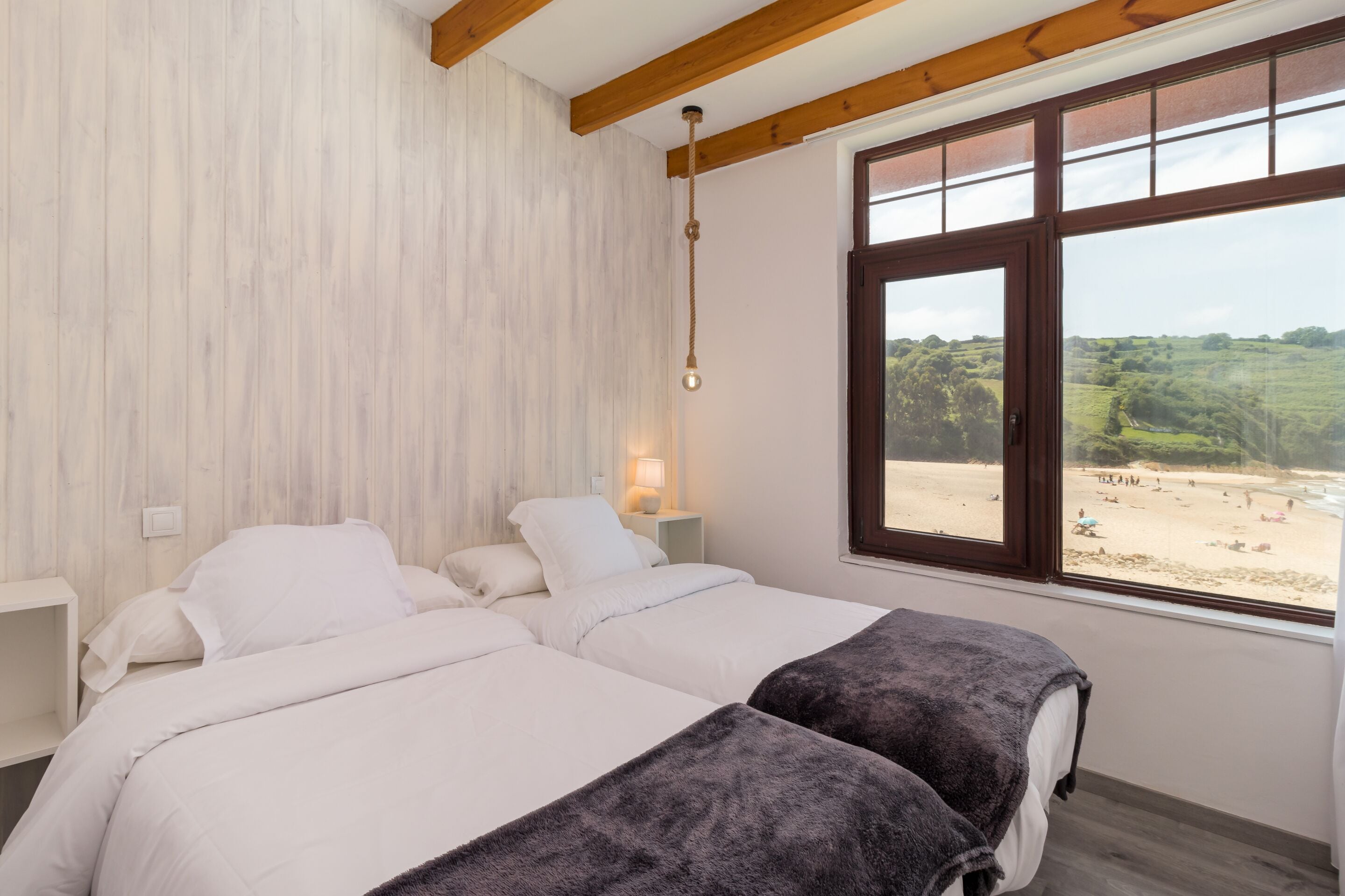 A bedroom with two beds, a wooden ceiling, a large window with a beach view, and a bedside lamp.