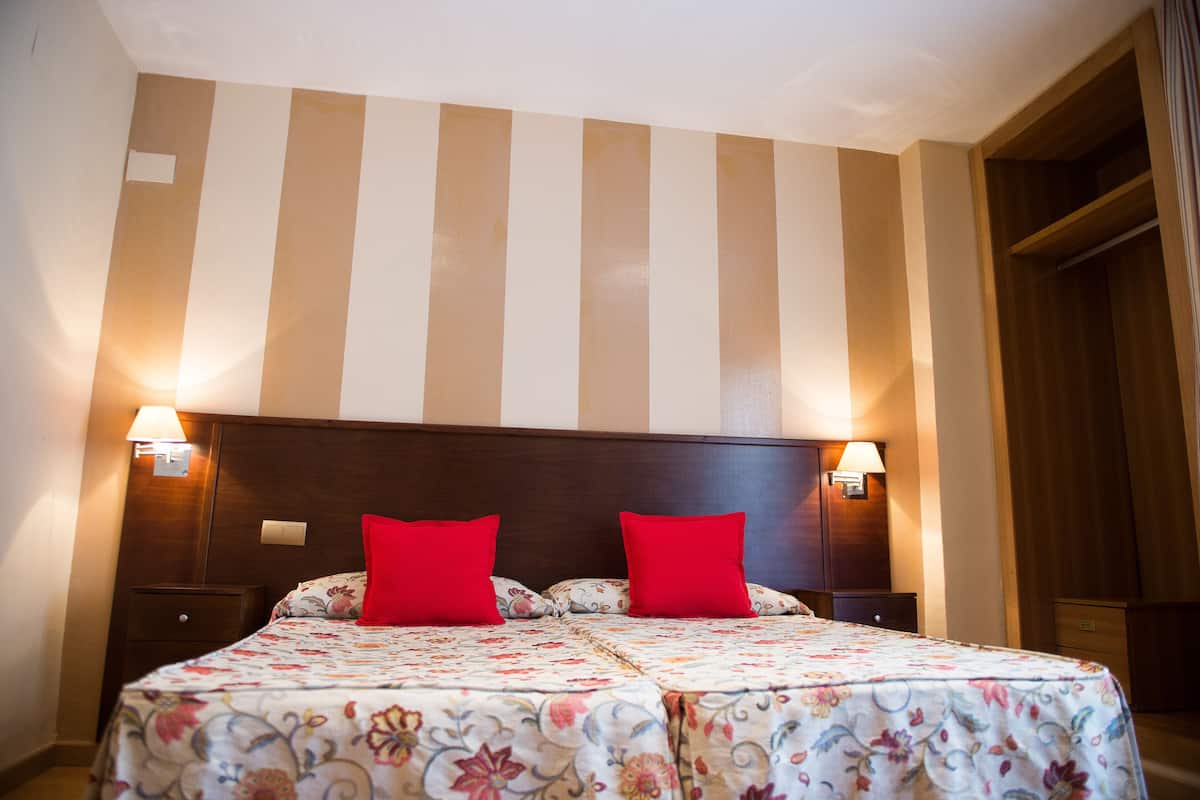 Un lit double avec une literie à motifs floraux et deux oreillers rouges, encadré par des tables de chevet sur lesquelles sont posées des lampes murales, dans une pièce dont le papier peint est rayé et dont les meubles sont en bois.