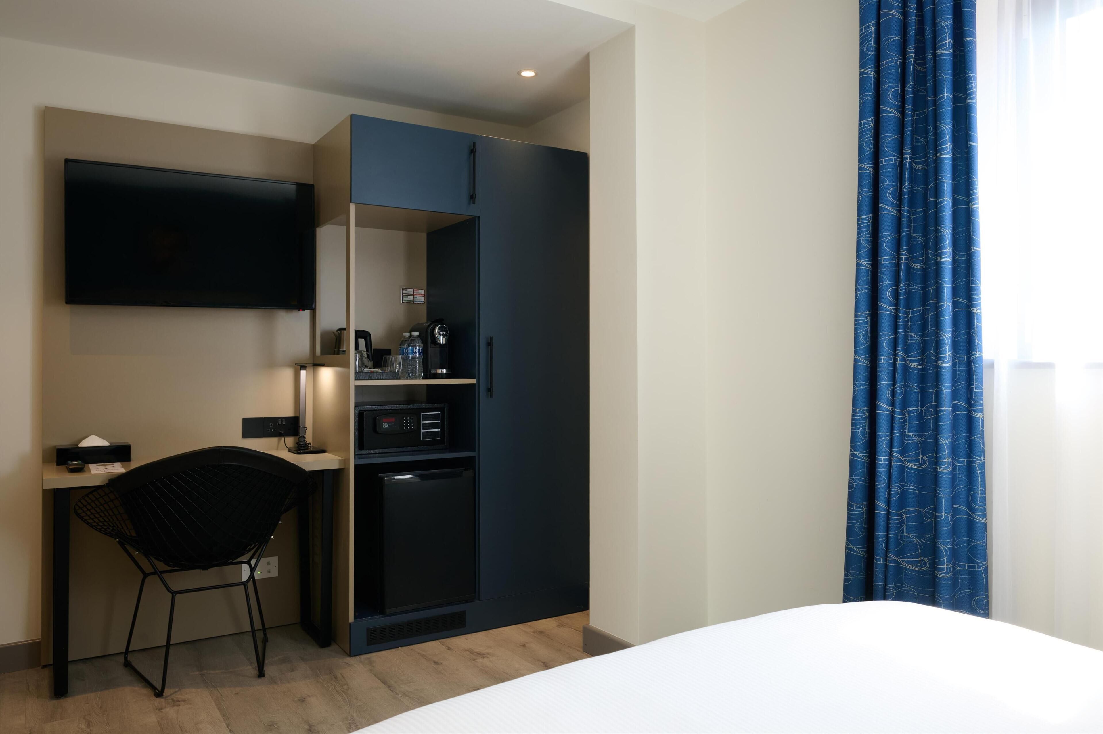 A hotel room with a bed, a desk with a chair, a TV, a mini-fridge, and a wardrobe.