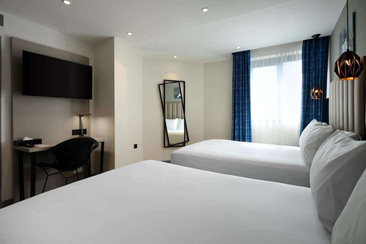 A hotel room with two beds, a desk, a chair, a TV, a mirror, and a window with curtains.