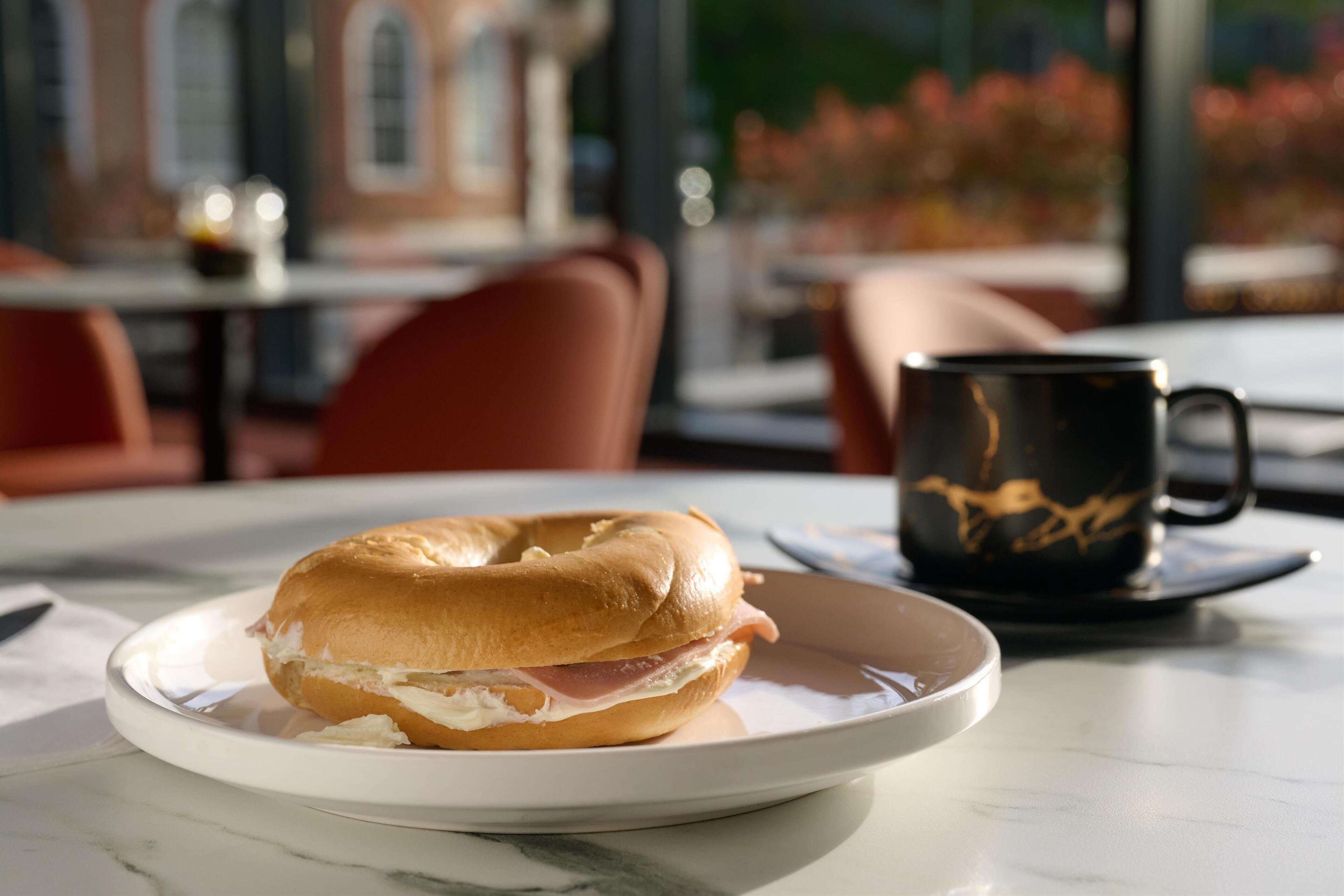 A breakfast setting with a bagel sandwich on a plate, a black mug, and a table with a view of a building and flowers through the window.