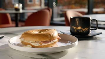 A breakfast setting with a bagel sandwich on a plate, a black mug, and a table with a view of a building and flowers through the window.