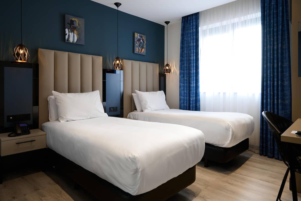 A hotel room with two beds, a desk, and a chair. There are artwork on the walls and a window with curtains.