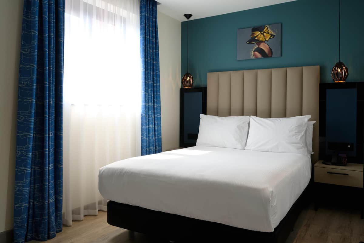 A hotel room with a large bed, two bedside tables, a headboard, a painting on the wall, and curtains.