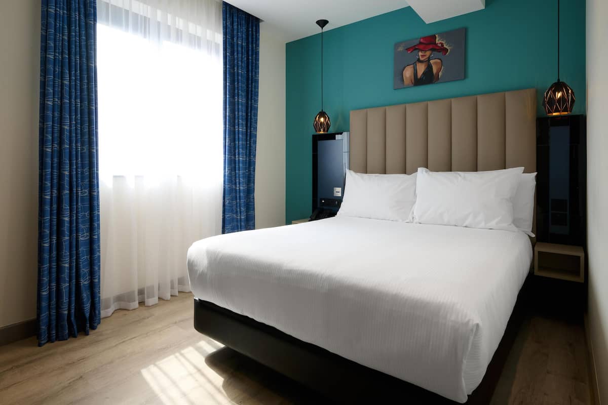 A modern hotel room with a large bed, a nightstand, a wall-mounted lamp, and a painting.