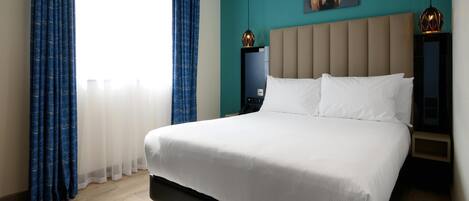 A modern hotel room with a large bed, a nightstand, a wall-mounted lamp, and a painting.