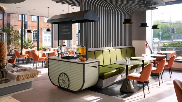 A modern cafe with a central seating area featuring green cushioned benches, surrounded by orange chairs and tables. A vintage-style counter with a bicycle and a fruit bowl is visible. - Spark by Hilton Luton (Luton)