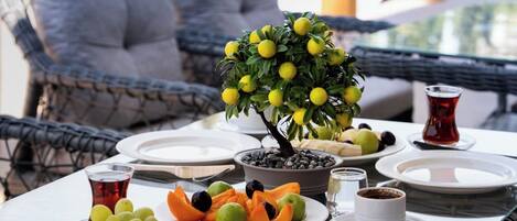 An outdoor dining area with wicker furniture, a table set with plates of fruit, a small tree, and a glass of red beverage.